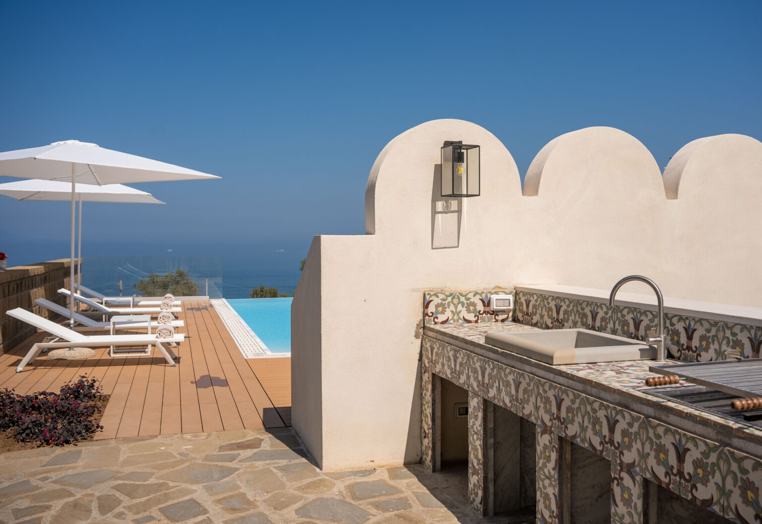 A luxurious outdoor space featuring a swimming pool, sun loungers, and a stylish outdoor kitchen area, with a clear blue sky and ocean view in the background. | Only on SopranoVillas