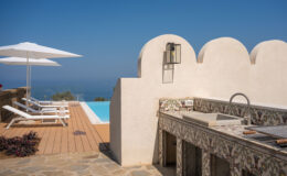 A luxurious outdoor space featuring a swimming pool, sun loungers, and a stylish outdoor kitchen area, with a clear blue sky and ocean view in the background.