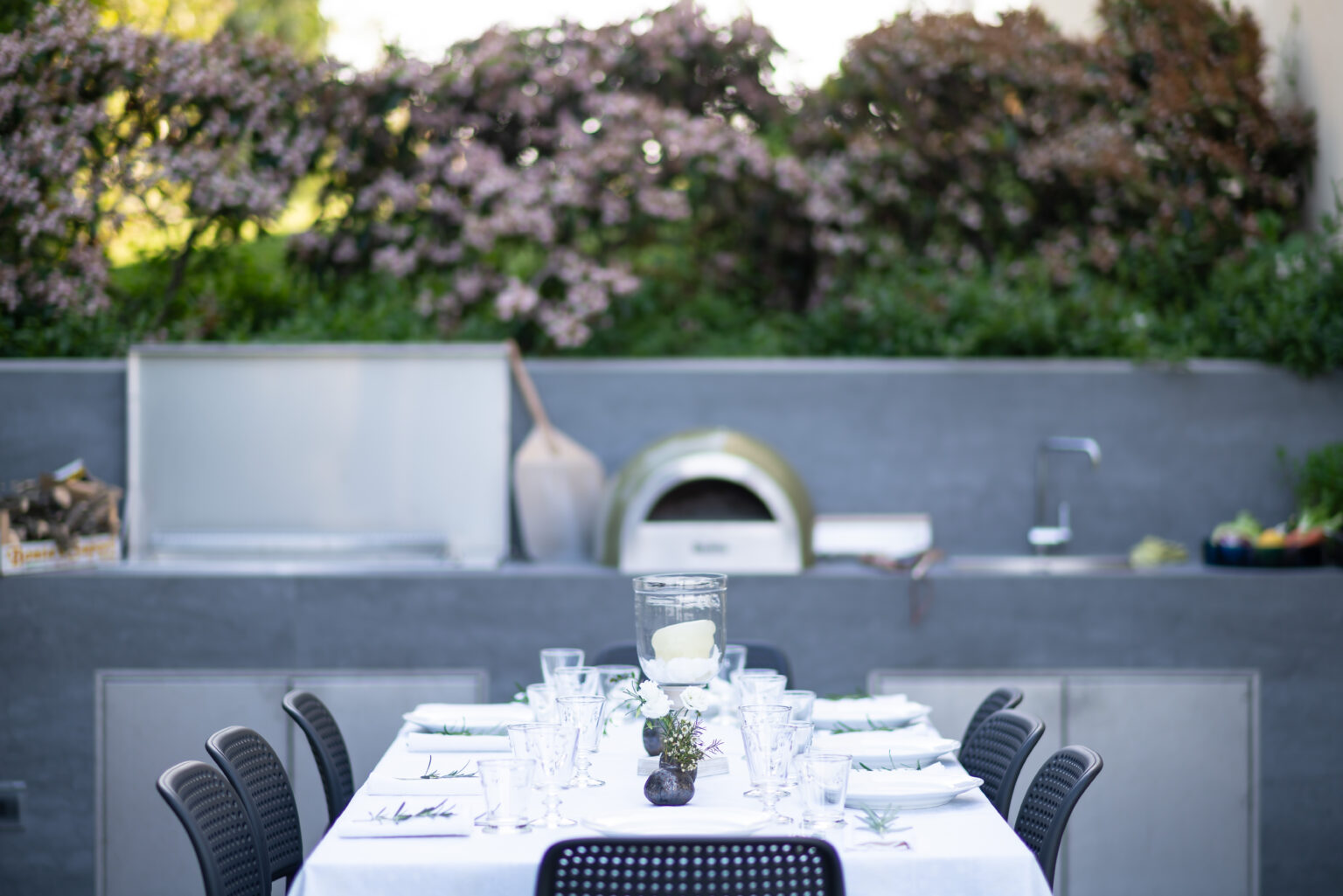 Outdoor dining setup featuring a long table with elegant place settings, surrounded by blooming flowers and a modern outdoor kitchen with a pizza oven. Ideal for al fresco gatherings and entertaining guests. | Only on SopranoVillas