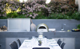 Outdoor dining setup featuring a long table with elegant place settings, surrounded by blooming flowers and a modern outdoor kitchen with a pizza oven. Ideal for al fresco gatherings and entertaining guests.