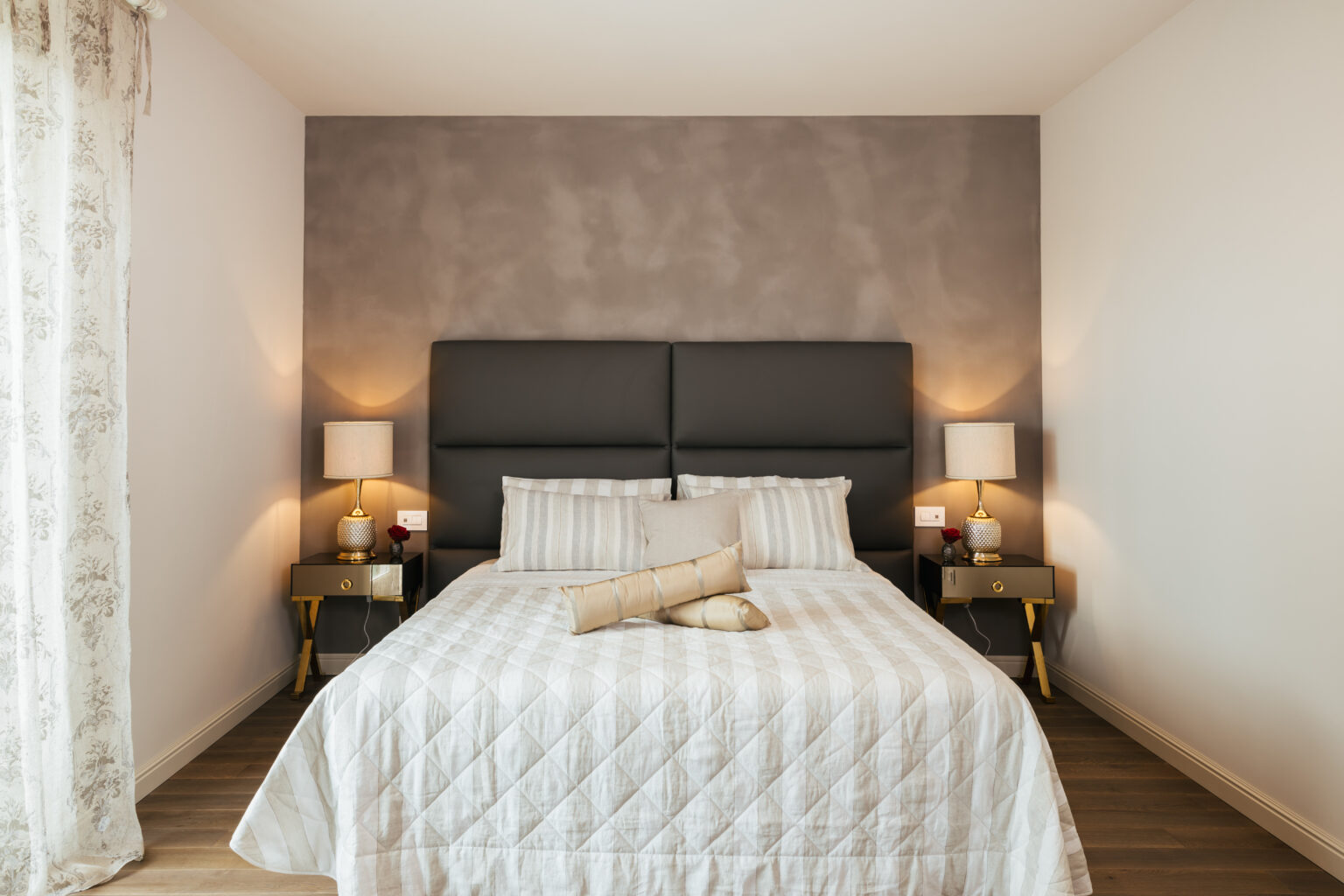 Modern bedroom design featuring a queen-sized bed with a gray upholstered headboard, white striped bedding, and decorative pillows. Two bedside tables with stylish lamps flank the bed, complemented by a neutral color palette and wooden flooring. Ideal for a cozy and elegant interior aesthetic. | Only on SopranoVillas