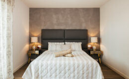 Modern bedroom design featuring a queen-sized bed with a gray upholstered headboard, white striped bedding, and decorative pillows. Two bedside tables with stylish lamps flank the bed, complemented by a neutral color palette and wooden flooring. Ideal for a cozy and elegant interior aesthetic.