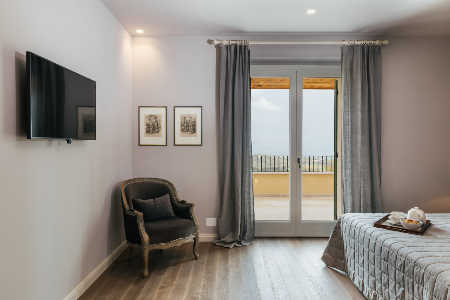 Modern bedroom interior featuring a comfortable armchair, a wall-mounted TV, and large glass doors leading to a balcony with outdoor views. The space is decorated with framed artwork and elegant curtains, creating a cozy and inviting atmosphere. | Only on SopranoVillas