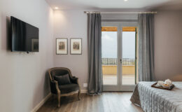 Modern bedroom interior featuring a comfortable armchair, a wall-mounted TV, and large glass doors leading to a balcony with outdoor views. The space is decorated with framed artwork and elegant curtains, creating a cozy and inviting atmosphere.