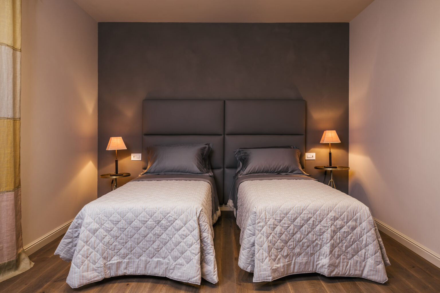 Modern hotel room featuring two single beds with gray upholstered headboards, soft lighting from bedside lamps, and a minimalist design. Ideal for travelers seeking comfort and style. | Only on SopranoVillas