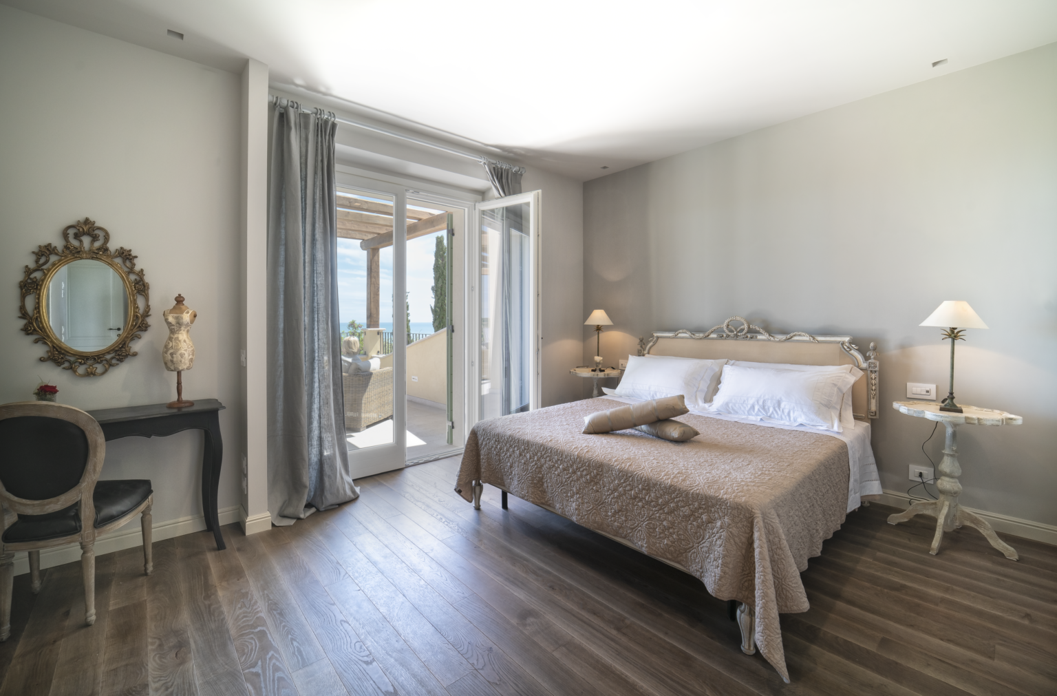 Spacious and elegantly decorated bedroom featuring a king-sized bed with neutral bedding, wooden flooring, large windows with balcony access, a vintage mirror, and stylish lamps, creating a serene and inviting atmosphere. | Only on SopranoVillas