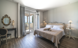 Spacious and elegantly decorated bedroom featuring a king-sized bed with neutral bedding, wooden flooring, large windows with balcony access, a vintage mirror, and stylish lamps, creating a serene and inviting atmosphere.