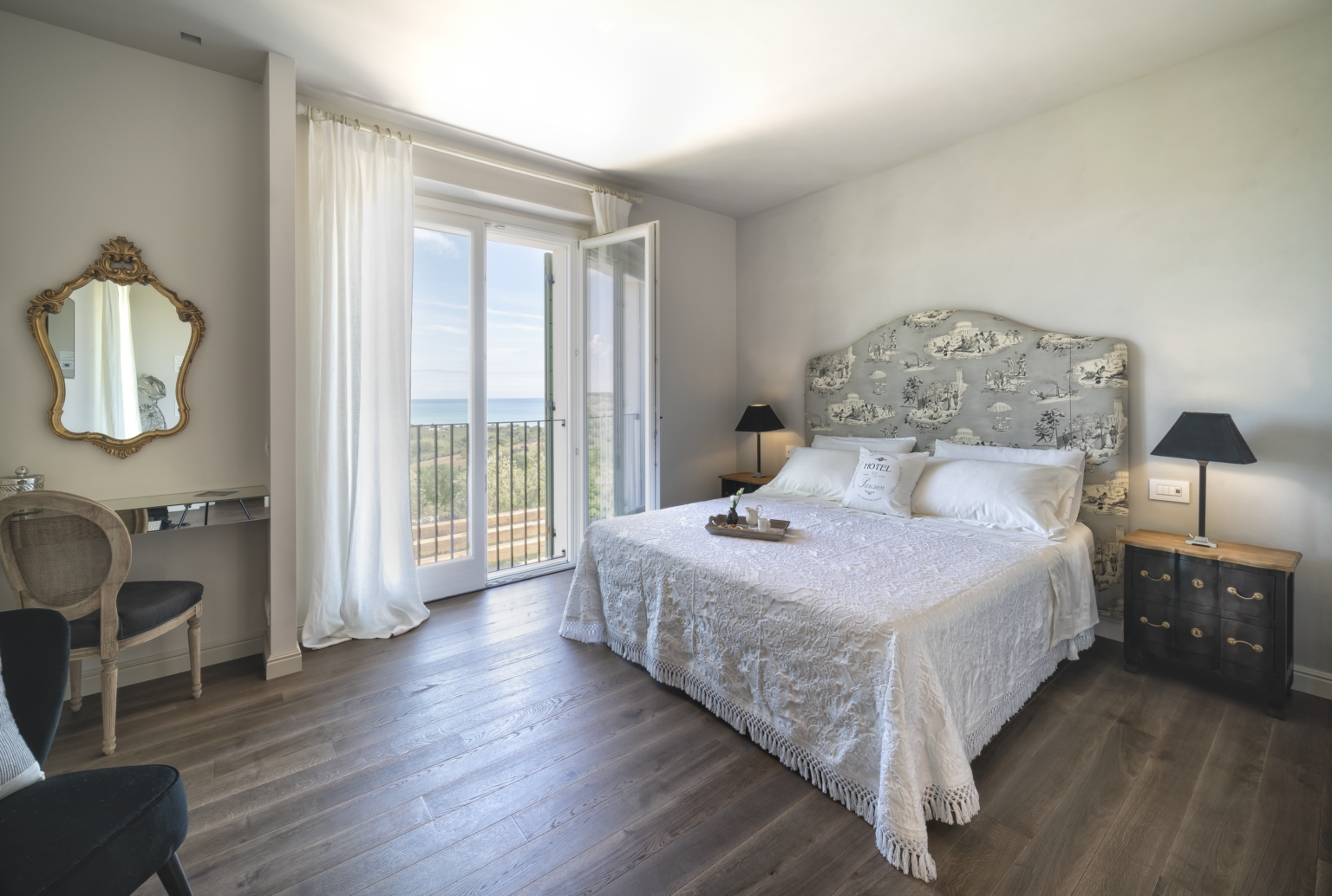 Cozy and elegant bedroom featuring a plush white bed with decorative headboard, large windows with scenic views, and stylish furnishings including a mirror and side tables, perfect for a relaxing getaway. | Only on SopranoVillas