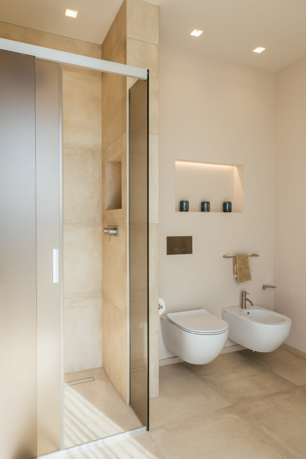 Modern bathroom interior featuring a glass shower enclosure, warm beige tiles, and minimalist decor. The design includes a recessed shelf with decorative candles, a towel rack, and a sleek faucet, emphasizing a clean and contemporary aesthetic. | Only on SopranoVillas