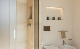 Modern bathroom interior featuring a glass shower enclosure, warm beige tiles, and minimalist decor. The design includes a recessed shelf with decorative candles, a towel rack, and a sleek faucet, emphasizing a clean and contemporary aesthetic.