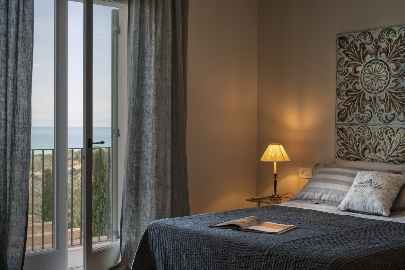 Cozy bedroom with a sea view, featuring a stylish bed with decorative pillows, a bedside lamp, and a window with sheer curtains. The room has a warm ambiance and elegant wall decor, perfect for relaxation. | Only on SopranoVillas