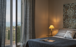 Cozy bedroom with a sea view, featuring a stylish bed with decorative pillows, a bedside lamp, and a window with sheer curtains. The room has a warm ambiance and elegant wall decor, perfect for relaxation.