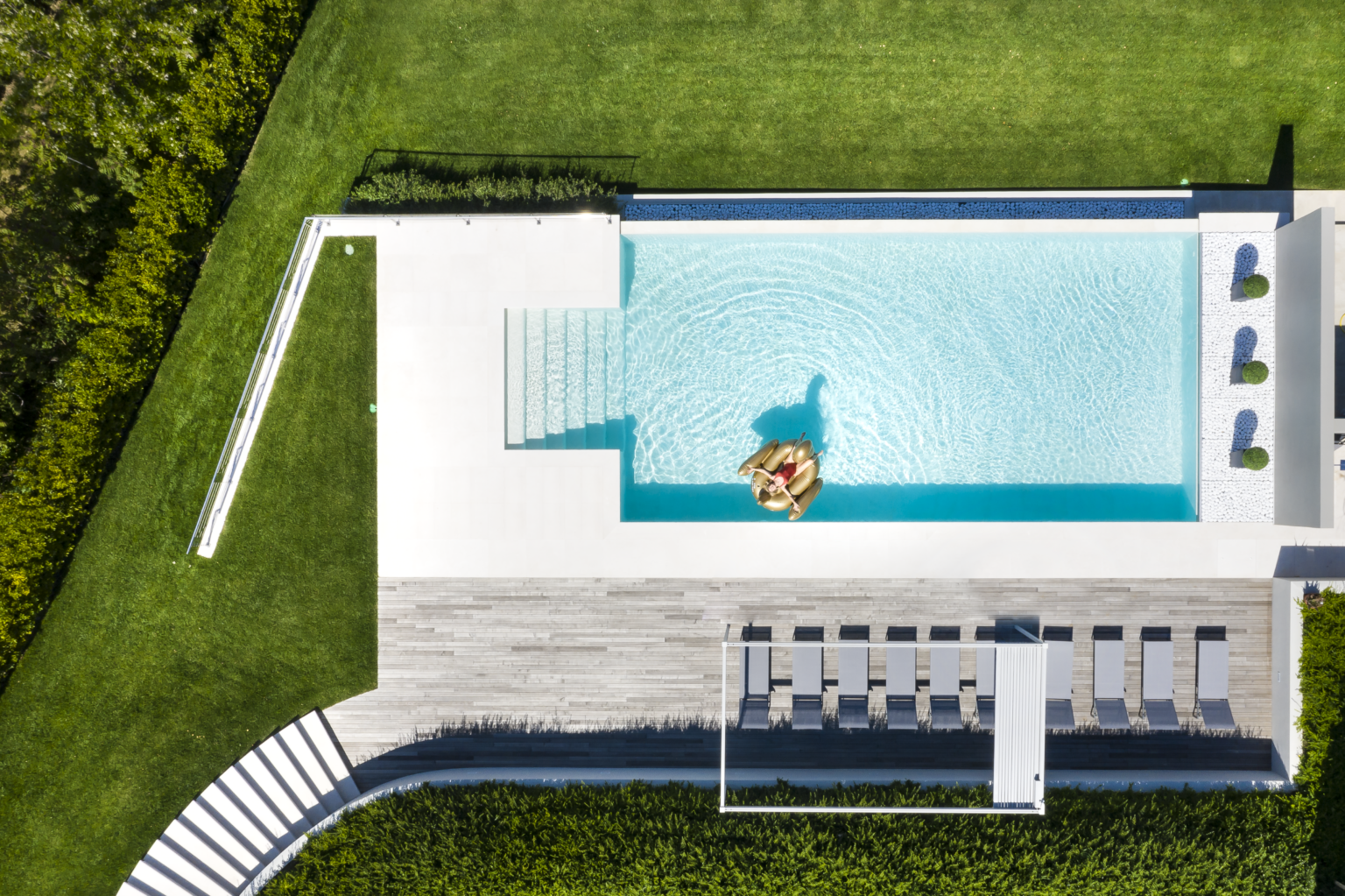 Aerial view of a modern backyard featuring a rectangular swimming pool with steps, surrounded by lush green grass and lounge chairs. The pool area is designed for relaxation, showcasing a stylish outdoor space perfect for summer gatherings. | Only on SopranoVillas