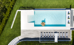 Aerial view of a modern backyard featuring a rectangular swimming pool with steps, surrounded by lush green grass and lounge chairs. The pool area is designed for relaxation, showcasing a stylish outdoor space perfect for summer gatherings.