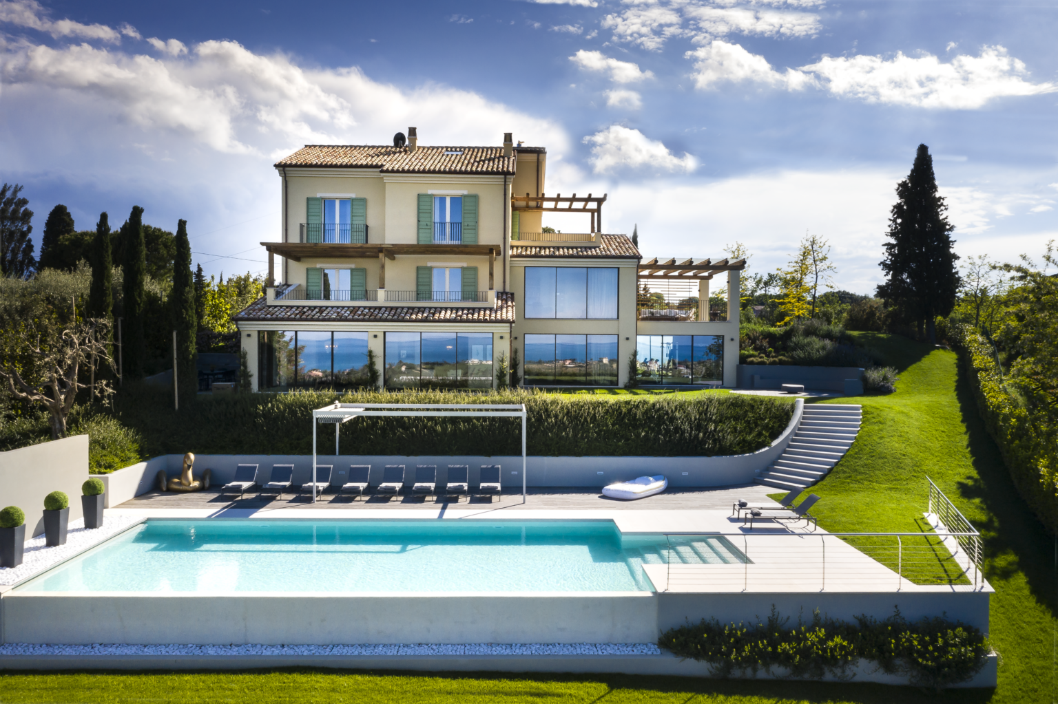 Luxurious villa featuring a modern design with large glass windows, surrounded by lush greenery and a serene pool area. The property includes a spacious terrace and relaxing outdoor seating, ideal for enjoying scenic views and outdoor living. | Only on SopranoVillas