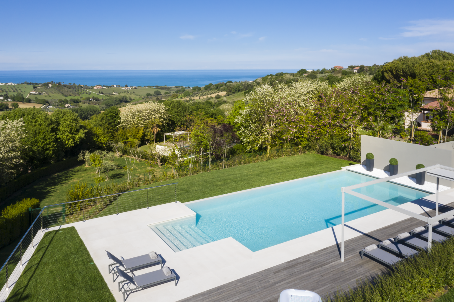Modern villa with a stunning infinity pool overlooking lush green hills and the ocean, featuring lounge chairs and a spacious deck. Ideal for luxury relaxation and scenic views. | Only on SopranoVillas