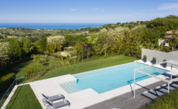 Modern villa with a stunning infinity pool overlooking lush green hills and the ocean, featuring lounge chairs and a spacious deck. Ideal for luxury relaxation and scenic views.