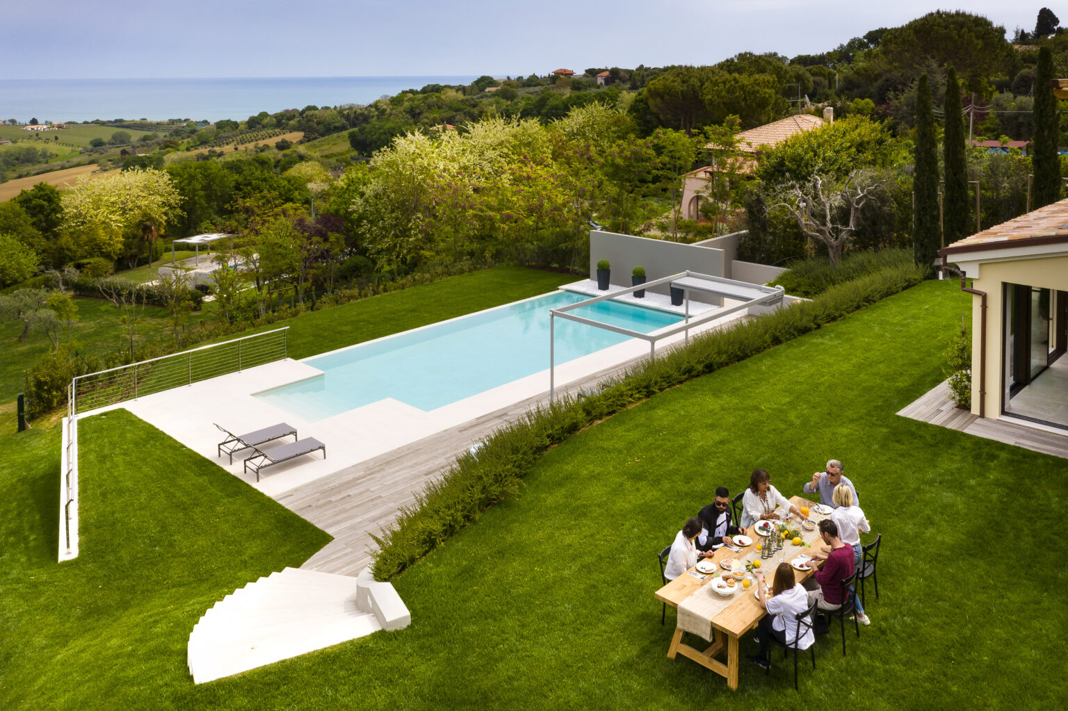 Aerial view of a modern villa with a swimming pool, surrounded by lush greenery and ocean views. A group of people enjoys a meal at a wooden dining table on the lawn, with lounge chairs nearby and landscaped gardens in the background. | Only on SopranoVillas