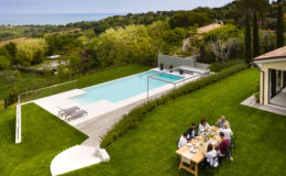 Aerial view of a modern villa with a swimming pool, surrounded by lush greenery and ocean views. A group of people enjoys a meal at a wooden dining table on the lawn, with lounge chairs nearby and landscaped gardens in the background.