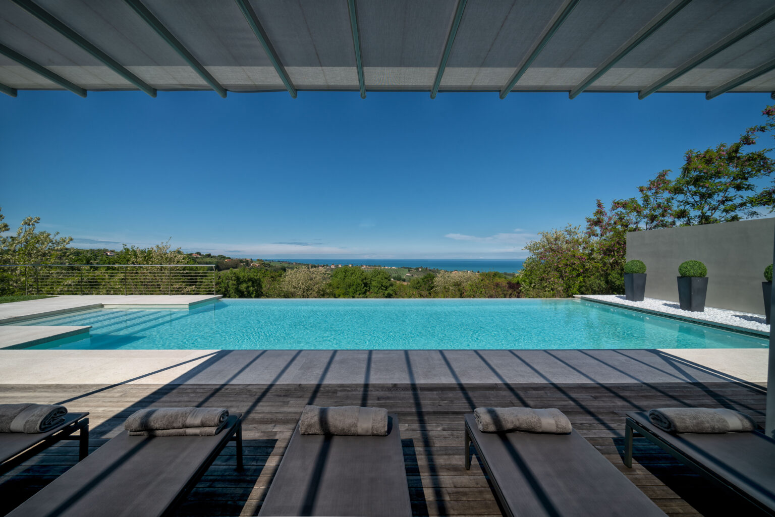 Modern outdoor pool area with lounge chairs, overlooking lush greenery and a distant ocean view, under a shaded pergola on a sunny day. | Only on SopranoVillas