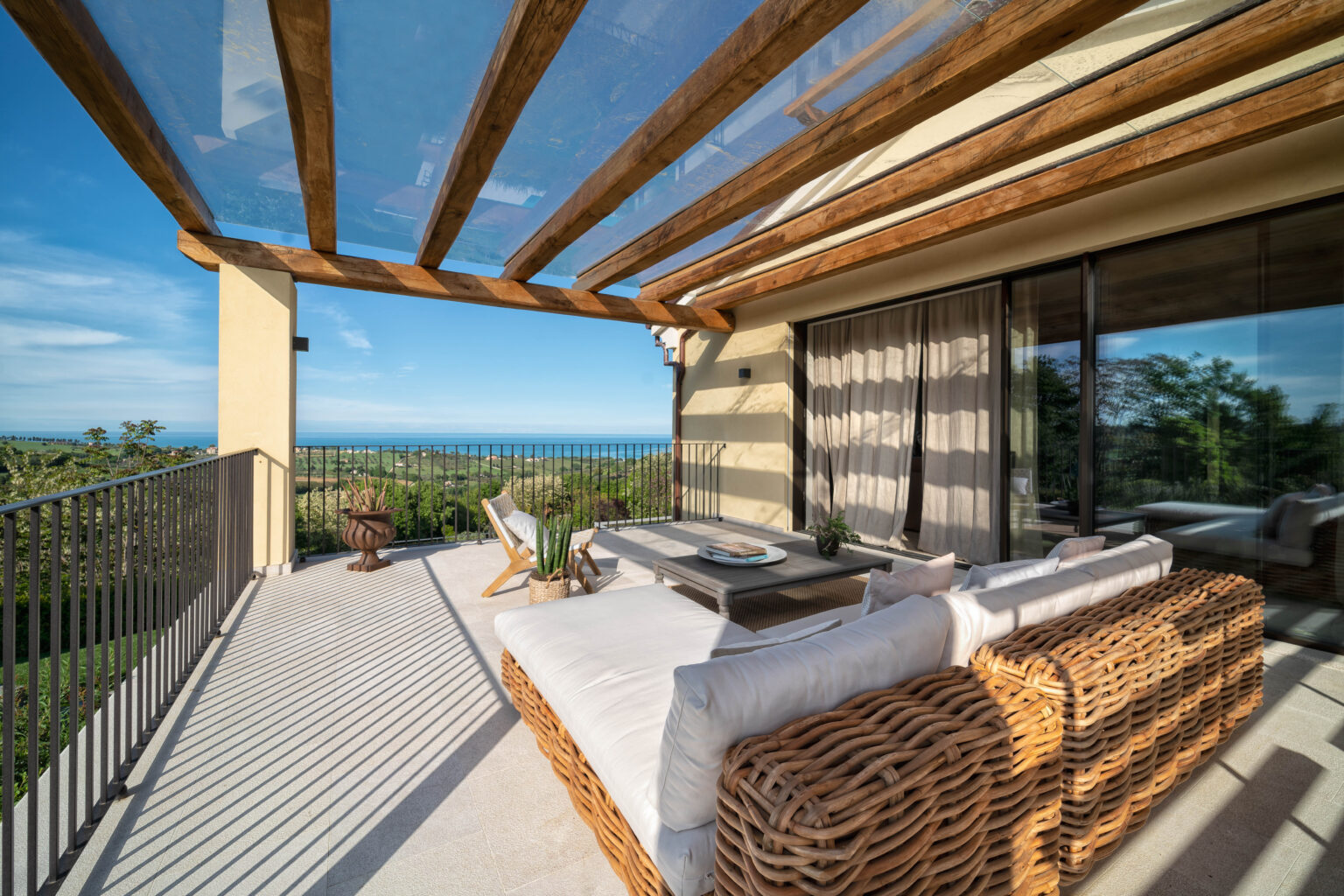 Stylish outdoor balcony with a panoramic ocean view, featuring a comfortable wicker sofa, a wooden coffee table, and potted plants under a wooden pergola. Bright blue sky and lush greenery enhance the serene atmosphere. | Only on SopranoVillas