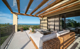 Stylish outdoor balcony with a panoramic ocean view, featuring a comfortable wicker sofa, a wooden coffee table, and potted plants under a wooden pergola. Bright blue sky and lush greenery enhance the serene atmosphere.