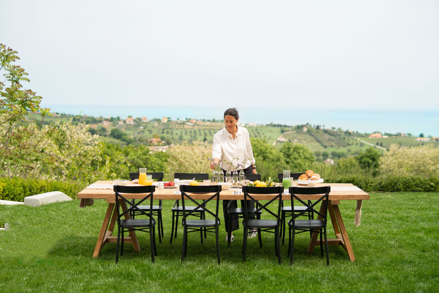Person preparing a vibrant outdoor dining setup on a wooden table with drinks and food, surrounded by a scenic landscape of hills and ocean in the background. Perfect for summer gatherings and alfresco dining. | Only on SopranoVillas