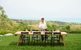 Person preparing a vibrant outdoor dining setup on a wooden table with drinks and food, surrounded by a scenic landscape of hills and ocean in the background. Perfect for summer gatherings and alfresco dining.