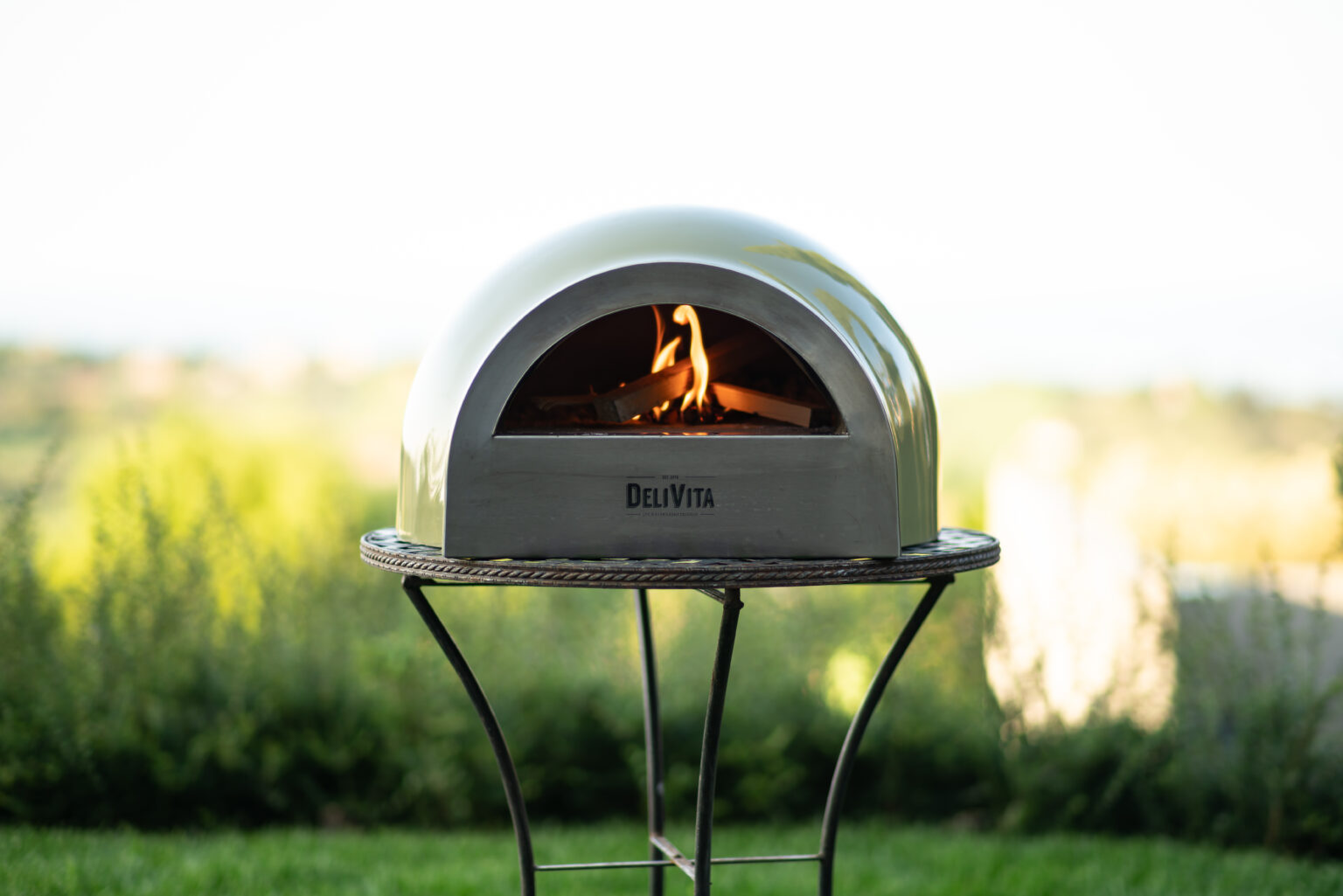 Outdoor wood-fired pizza oven with a sleek design, featuring a visible flame, set against a lush green landscape. Ideal for backyard cooking and entertaining. | Only on SopranoVillas
