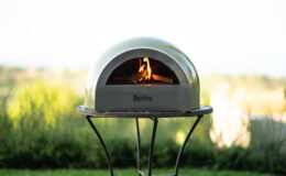 Outdoor wood-fired pizza oven with a sleek design, featuring a visible flame, set against a lush green landscape. Ideal for backyard cooking and entertaining.