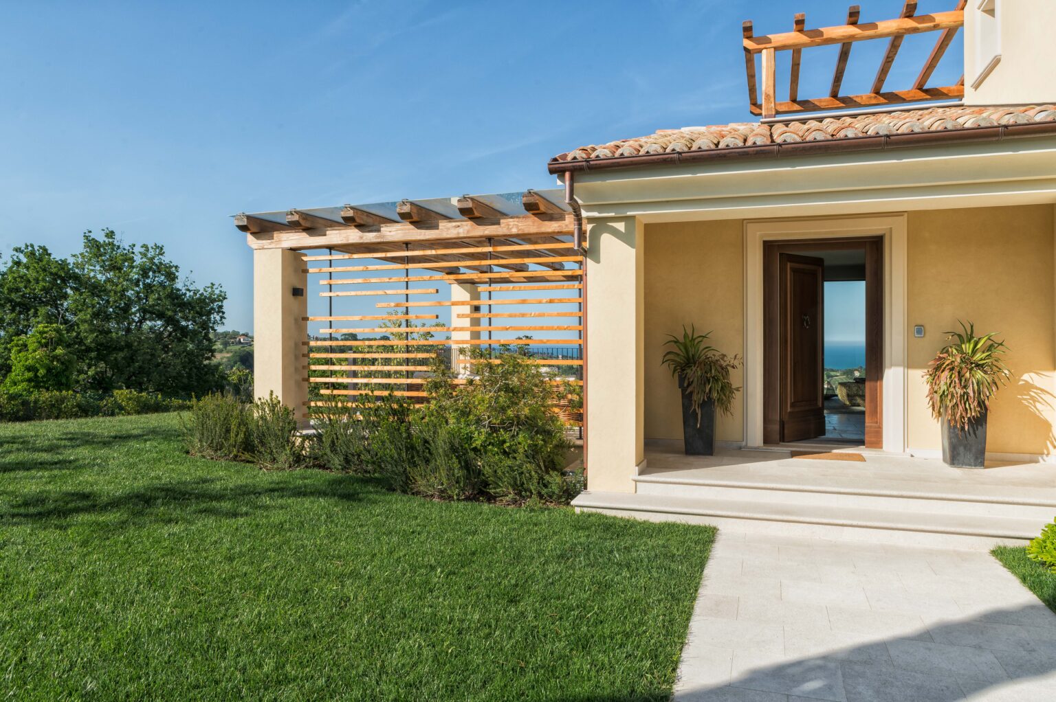 Modern home exterior with a spacious porch, lush green lawn, and wooden pergola, showcasing a scenic view of the surrounding landscape. Ideal for outdoor relaxation and entertaining. | Only on SopranoVillas