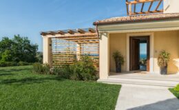 Modern home exterior with a spacious porch, lush green lawn, and wooden pergola, showcasing a scenic view of the surrounding landscape. Ideal for outdoor relaxation and entertaining.