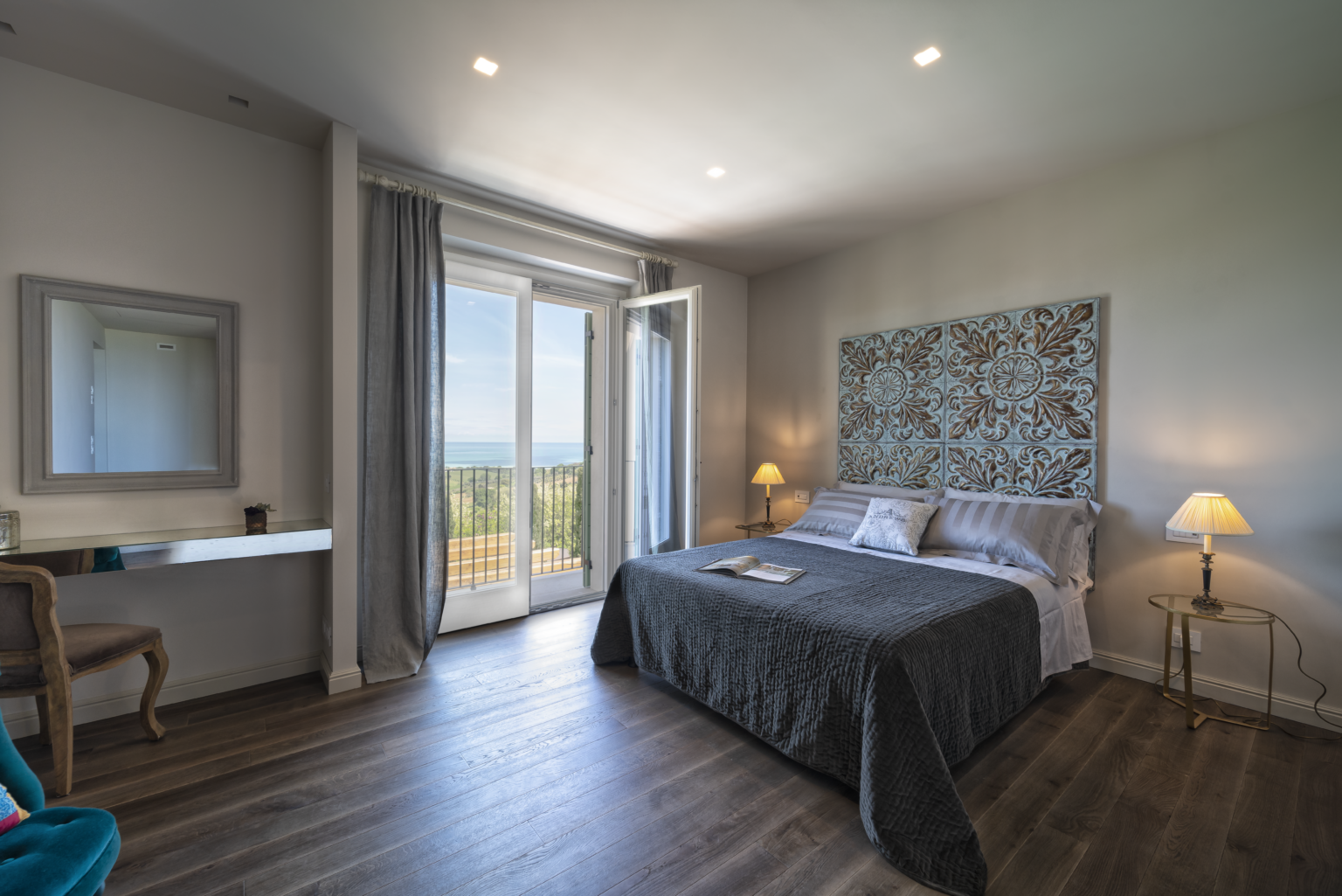 Modern bedroom with a large bed, stylish decor, and sliding glass doors leading to a balcony with scenic views. Features include a bedside lamp, a decorative wall panel, and a workspace with a mirror. The room has wooden flooring and natural light. | Only on SopranoVillas
