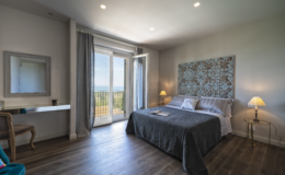 Modern bedroom with a large bed, stylish decor, and sliding glass doors leading to a balcony with scenic views. Features include a bedside lamp, a decorative wall panel, and a workspace with a mirror. The room has wooden flooring and natural light.