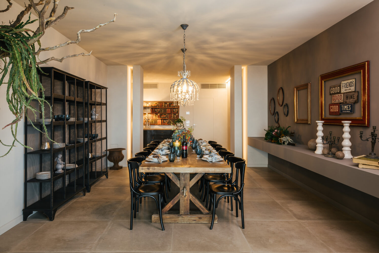Modern dining room featuring a wooden table set for a meal, surrounded by black chairs, with an elegant chandelier overhead. A stylish bookshelf and decorative elements add to the contemporary aesthetic. | Only on SopranoVillas