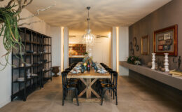 Modern dining room featuring a wooden table set for a meal, surrounded by black chairs, with an elegant chandelier overhead. A stylish bookshelf and decorative elements add to the contemporary aesthetic.