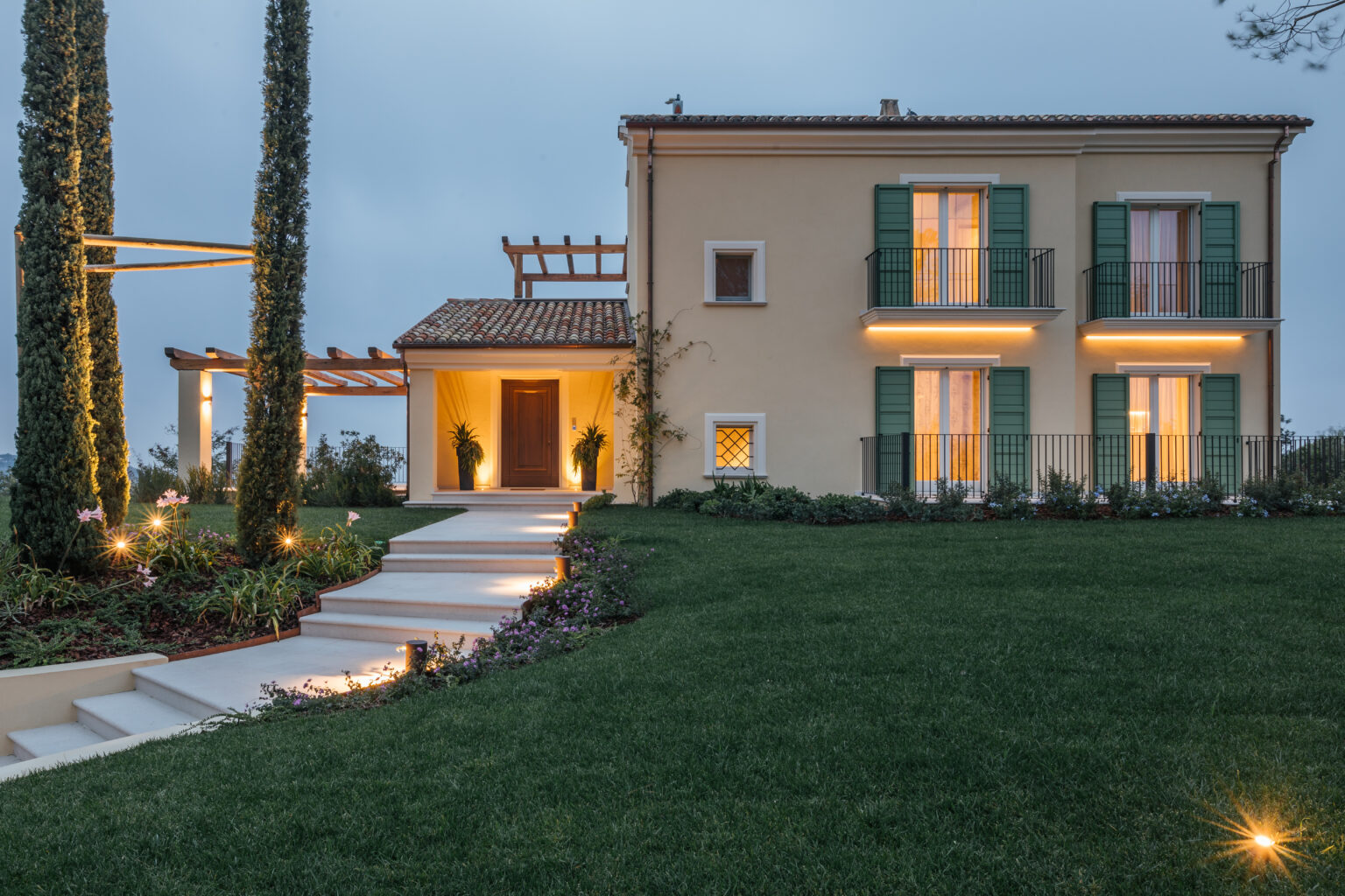 Elegant two-story house with green shutters, surrounded by landscaped gardens and illuminated pathways, showcasing modern architecture against a twilight sky. | Only on SopranoVillas