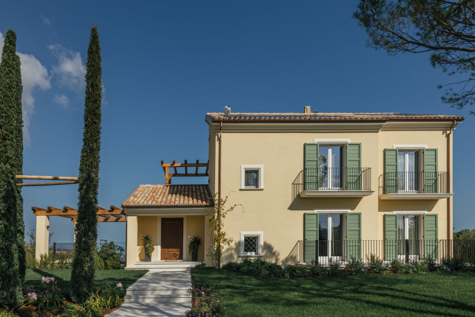 Two-story yellow house with green shutters, surrounded by landscaped gardens and tall cypress trees, under a clear blue sky. | Only on SopranoVillas