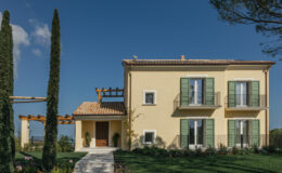 Two-story yellow house with green shutters, surrounded by landscaped gardens and tall cypress trees, under a clear blue sky.