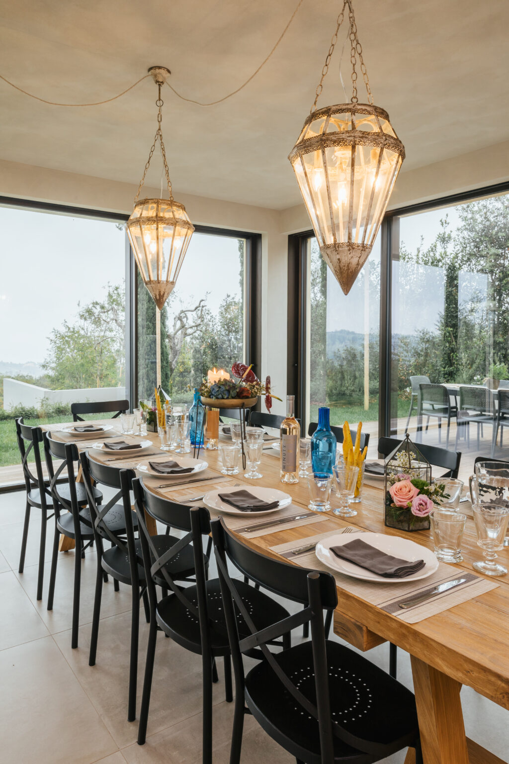 Elegant dining room setting featuring a long wooden table adorned with tableware, glassware, and floral arrangements, illuminated by stylish pendant lights. Large windows provide a scenic view of the outdoors, enhancing the inviting atmosphere. | Only on SopranoVillas