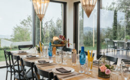 Elegant dining room setting featuring a long wooden table adorned with tableware, glassware, and floral arrangements, illuminated by stylish pendant lights. Large windows provide a scenic view of the outdoors, enhancing the inviting atmosphere.