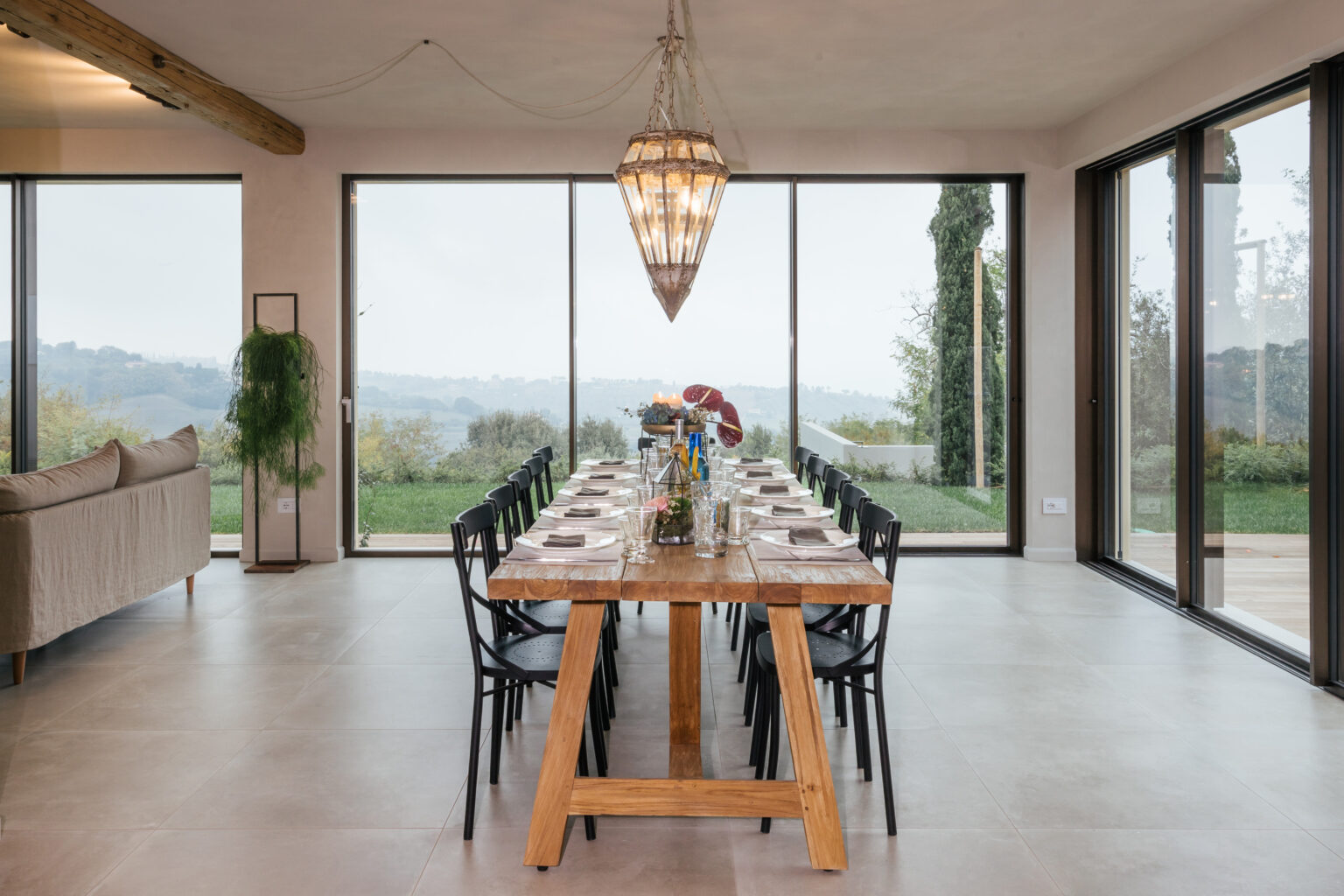 Modern dining room featuring a long wooden table set for a meal, surrounded by black chairs. Large windows provide a scenic view of the outdoors, with greenery visible in the background. The space is illuminated by a stylish chandelier, creating an inviting atmosphere. | Only on SopranoVillas