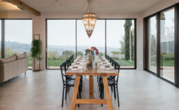Modern dining room featuring a long wooden table set for a meal, surrounded by black chairs. Large windows provide a scenic view of the outdoors, with greenery visible in the background. The space is illuminated by a stylish chandelier, creating an inviting atmosphere.
