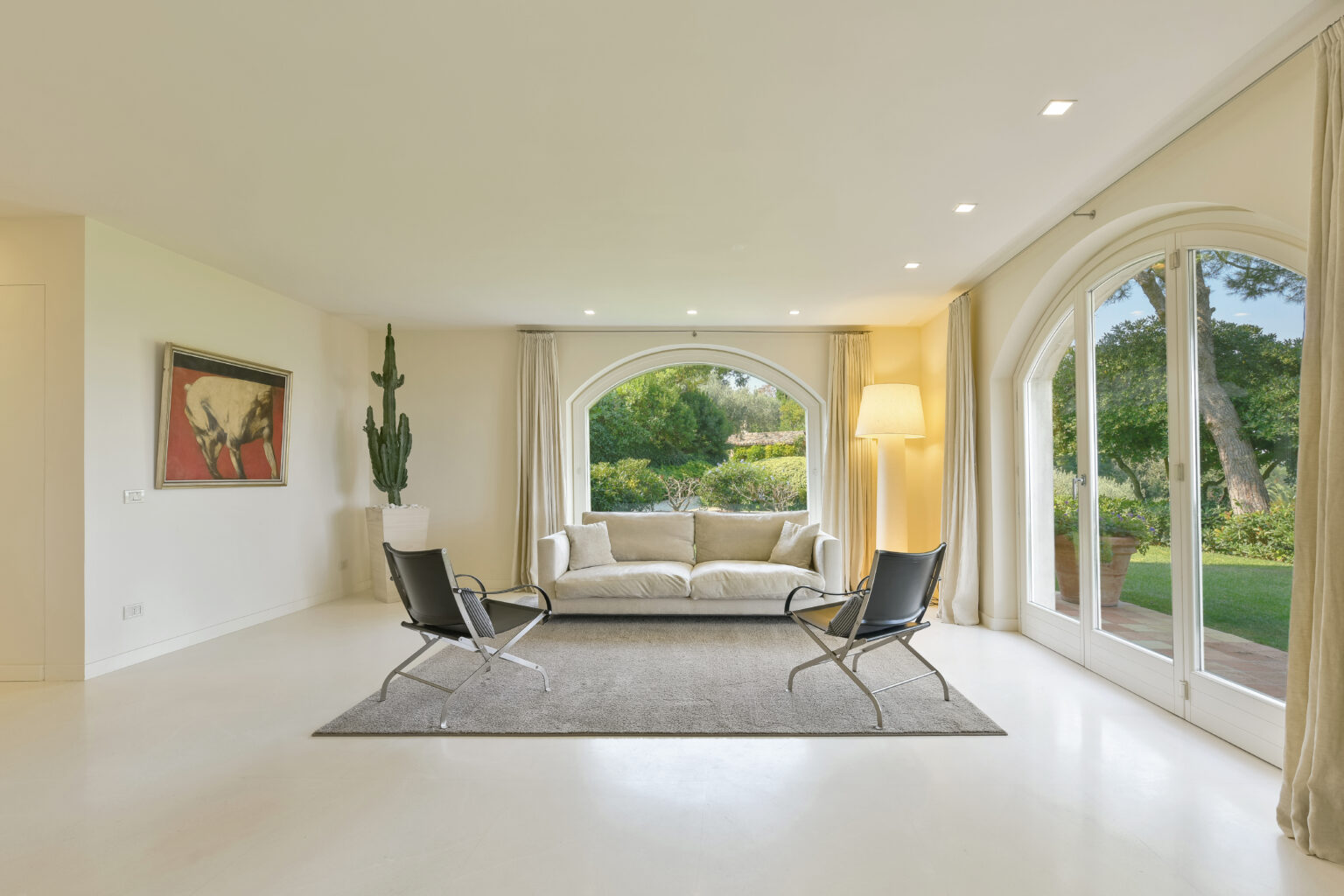 Modern living room with large arched windows, featuring a comfortable beige sofa, two black chairs, and a decorative rug, surrounded by greenery for a bright and inviting atmosphere. | Only on SopranoVillas