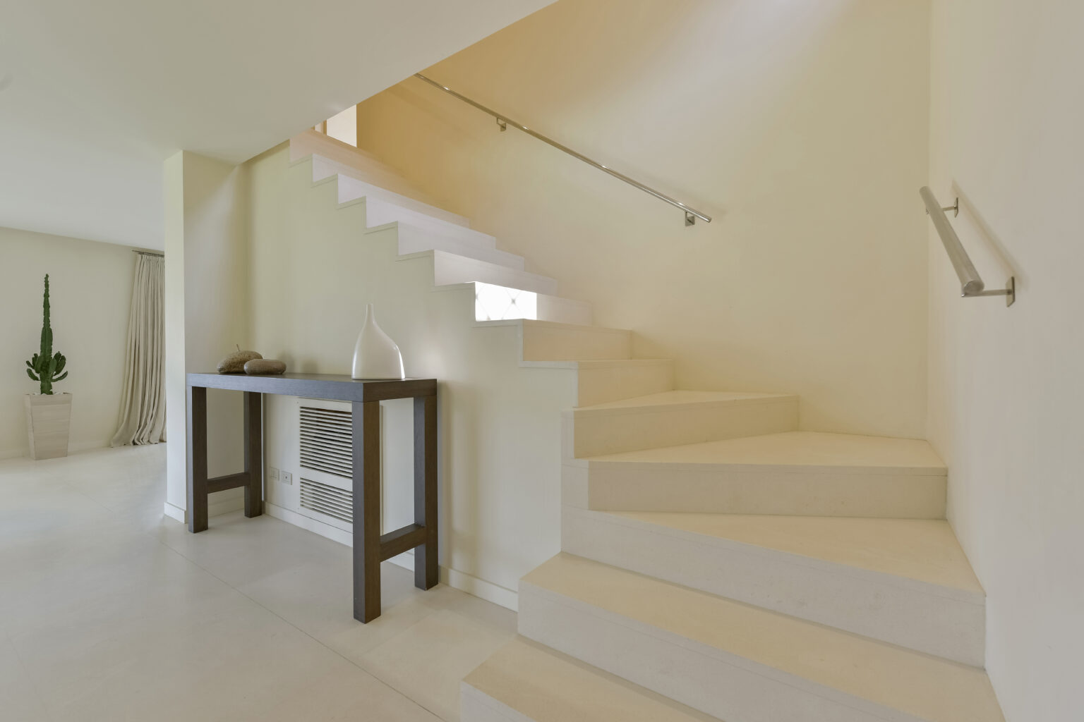 Modern staircase design in a bright interior, featuring white steps, a sleek handrail, and a minimalist console table with decorative items. A potted cactus adds a touch of greenery to the space. | Only on SopranoVillas