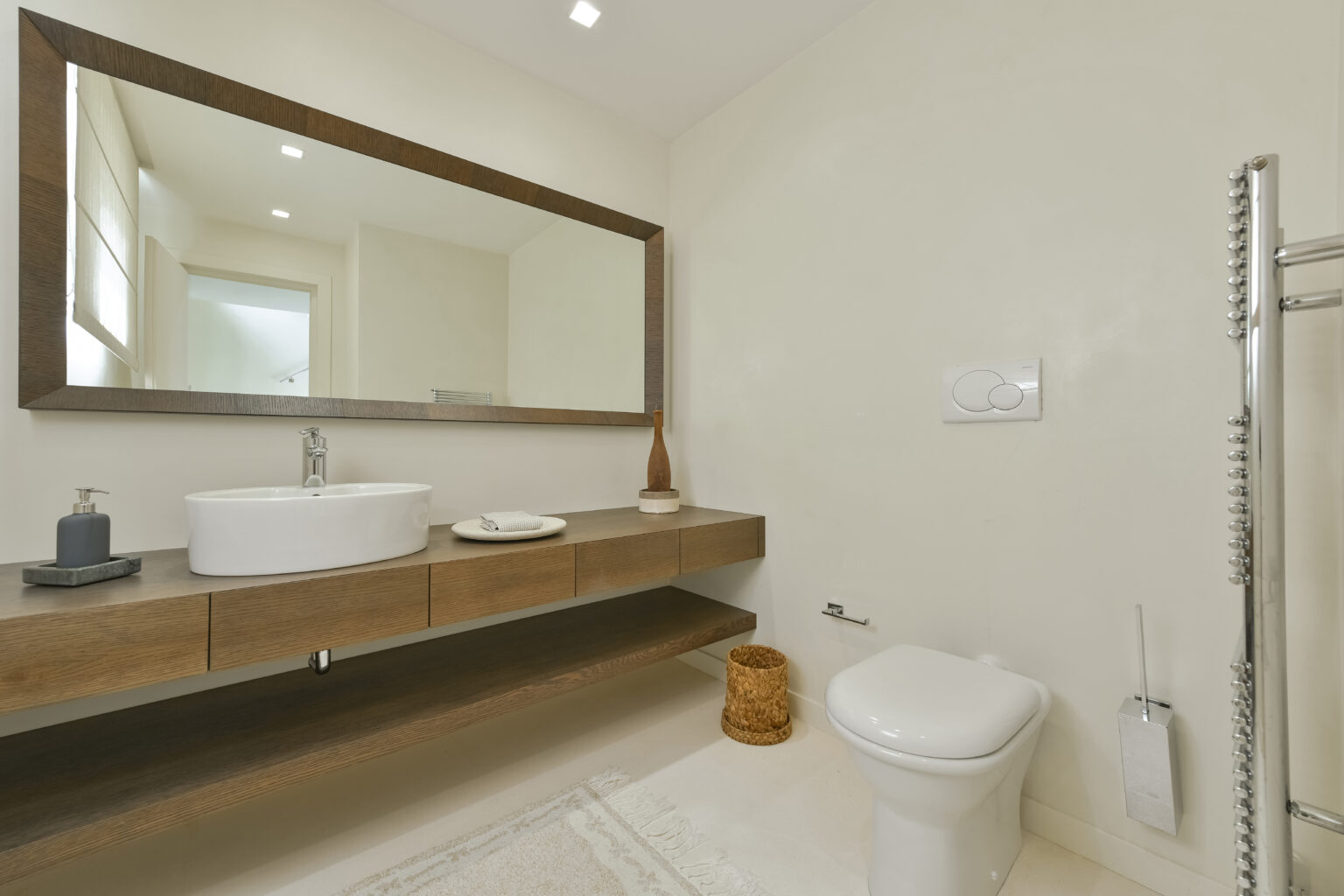 Modern bathroom interior featuring a minimalist design with a floating wooden vanity, oval sink, large mirror, and neutral color palette. The space includes a toilet, decorative storage basket, and stylish accessories, emphasizing a clean and contemporary aesthetic. | Only on SopranoVillas