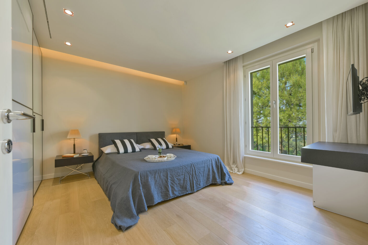 Modern bedroom featuring a gray bedspread, striped pillows, a nightstand with a lamp, and large windows with a view of greenery, complemented by warm lighting and wooden flooring. | Only on SopranoVillas