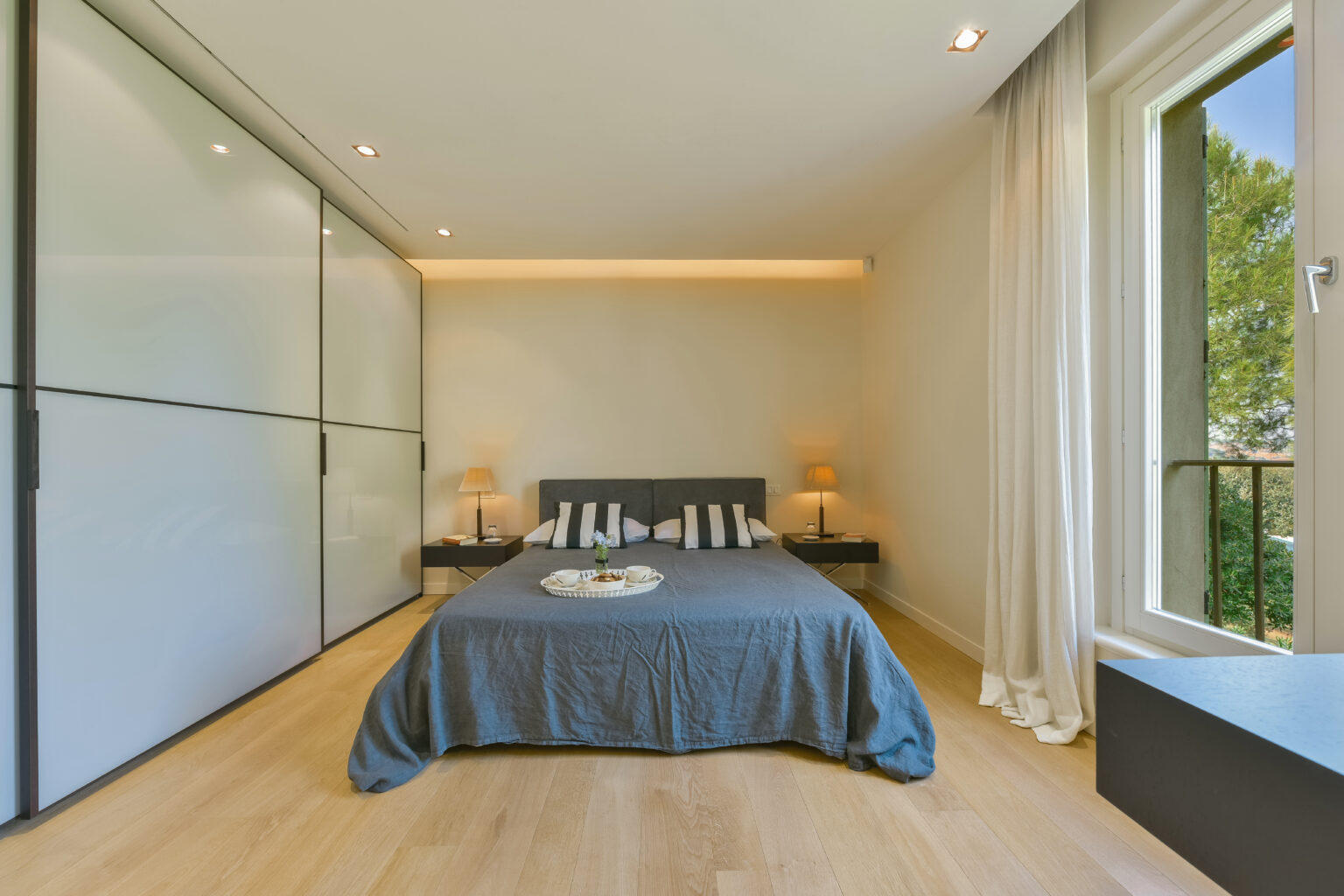 Modern bedroom featuring a gray bedspread, striped pillows, and minimalist decor. The room includes sleek sliding wardrobe doors, two bedside lamps, and a large window with a view of greenery, enhancing natural light and spaciousness. | Only on SopranoVillas