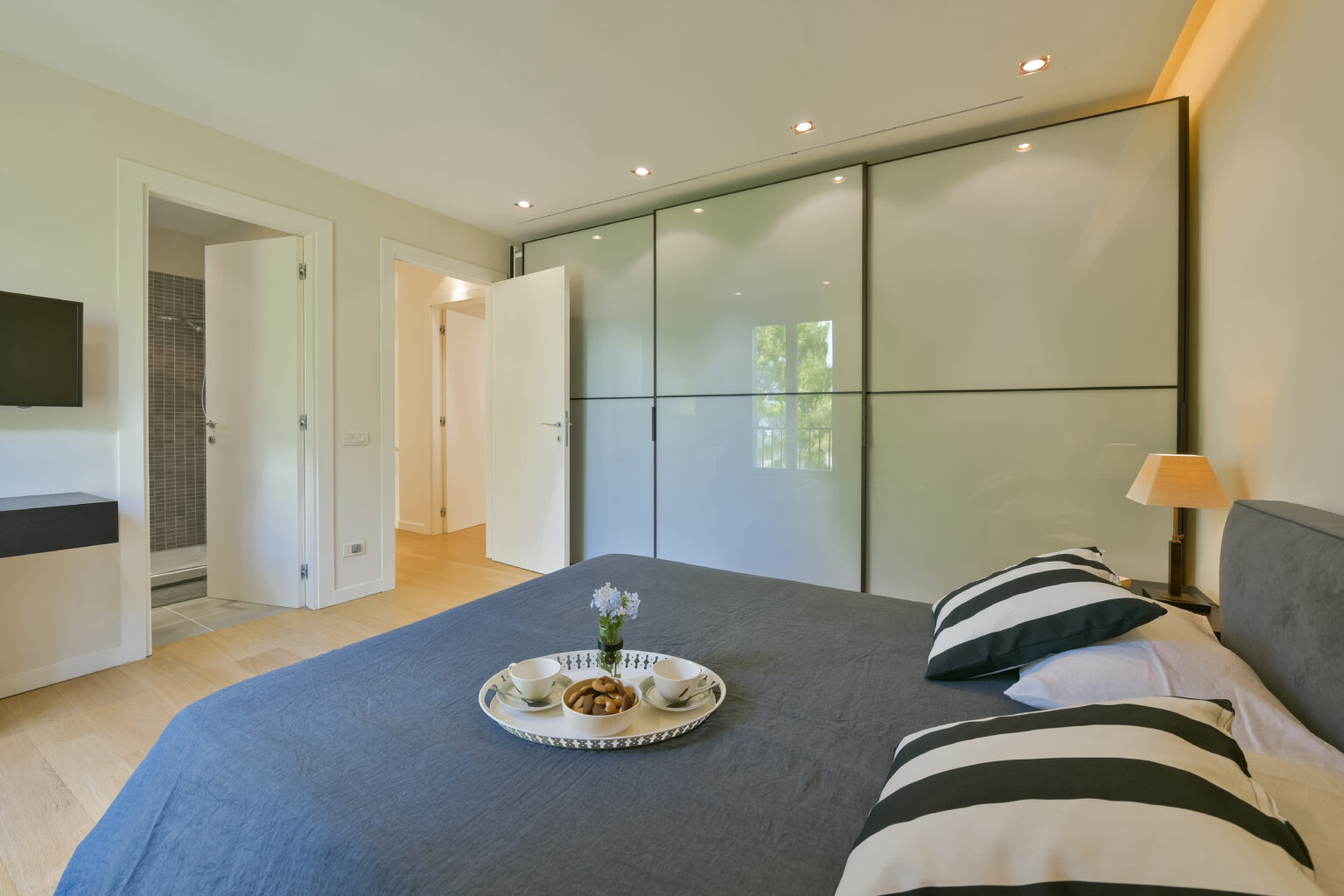Modern bedroom featuring a gray bedspread, decorative pillows, and a stylish tray with snacks on the bed. The room includes built-in wardrobes, soft lighting, and a wall-mounted TV, creating a cozy and inviting atmosphere. | Only on SopranoVillas