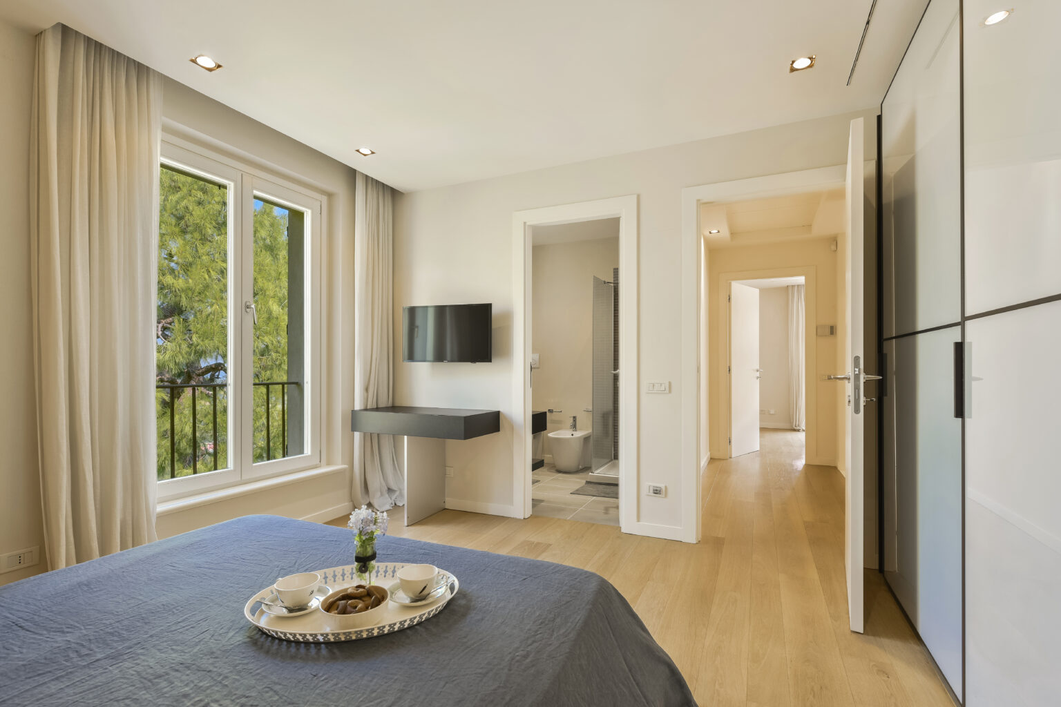 Modern bedroom featuring a cozy bed with a gray blanket, a small table with a decorative tray, and large windows providing natural light. The room includes a wall-mounted TV and leads to a bathroom, showcasing contemporary design and elegant decor. | Only on SopranoVillas